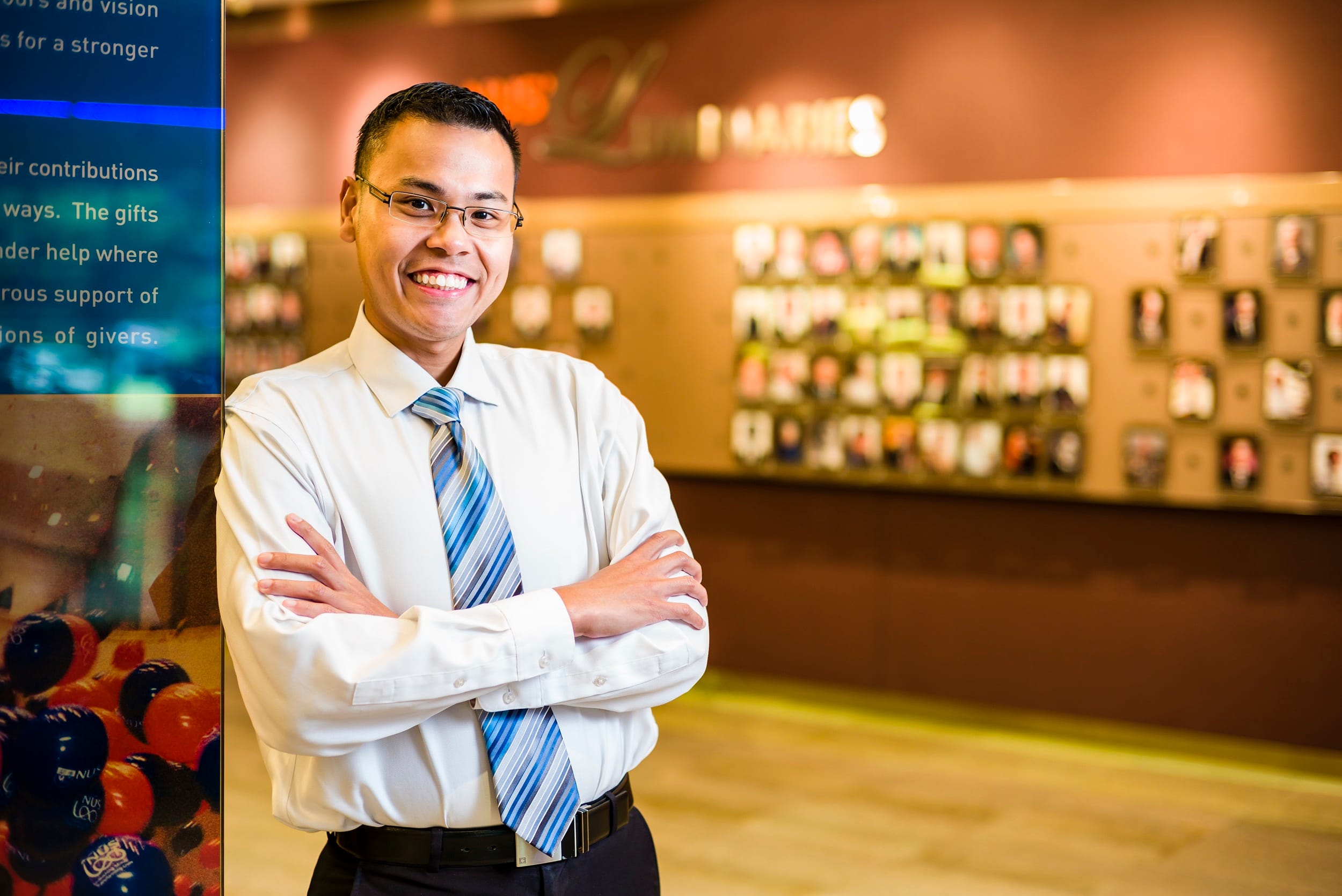 Smiling man with glasses in a white shirt and blue striped tie, arms crossed, standing in front of a photo display wall.
